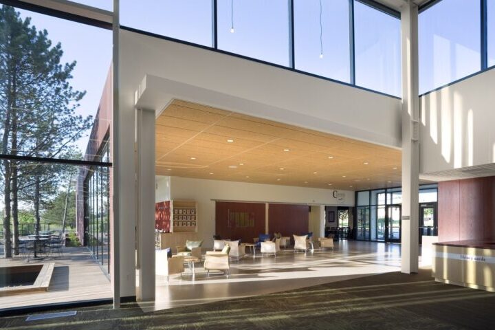 Interior modern building entry lobby with high ceiling and tall windows. Lots of natural light streams in and lounge chairs are clustered in an adjoining room.