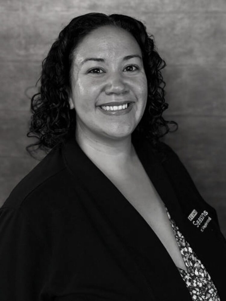 Black and white portrait of Gina Goldsmith. She wears a dark cardigan sweater over a blouse with a floral print.