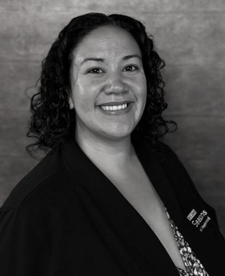 Black and white portrait of Gina Goldsmith. She wears a dark cardigan sweater over a blouse with a floral print.
