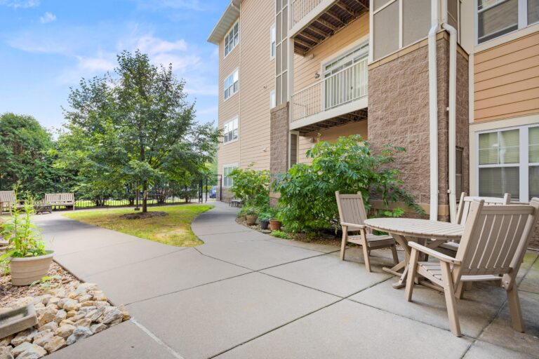 Sunny outdoor patio with tables and chairs next to a modern building and green trees.