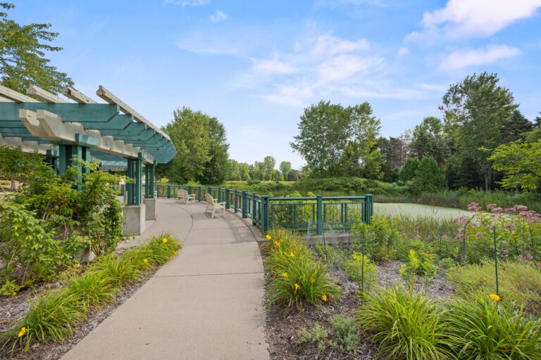 Curved pathway with a pergola and railing, surrounded by greenery and flowers, under a blue sky.
