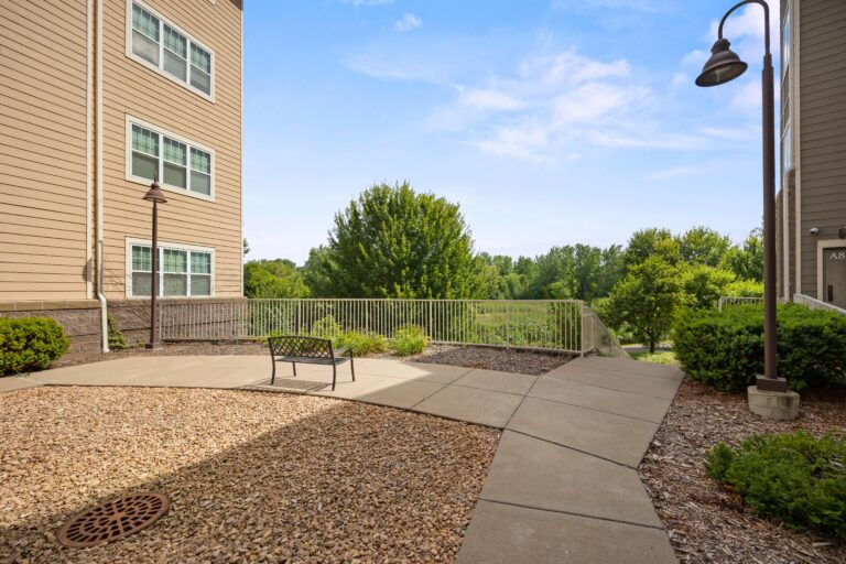 Outdoor seating area with a bench, surrounded by rocks and greenery, near apartment buildings under a blue sky.