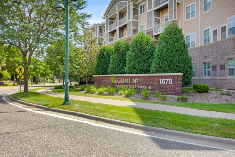 Entrance sign for Ecumen Seasons at Maplewood, beside a sidewalk, trees, and an apartment building.