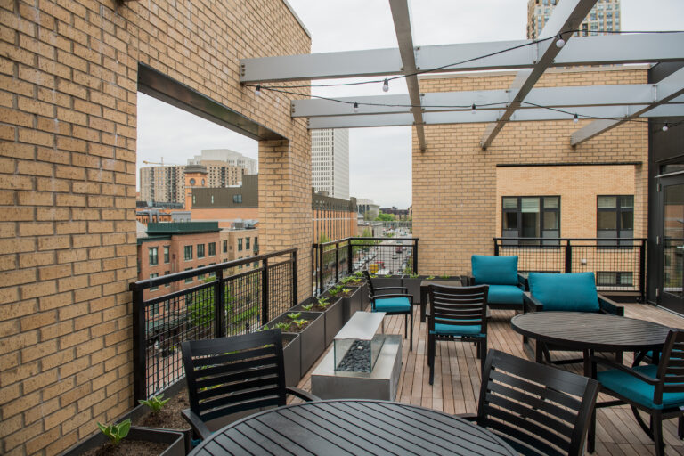 Modern rooftop patio with outdoor seating, a fire pit, potted plants, and city buildings in the background.