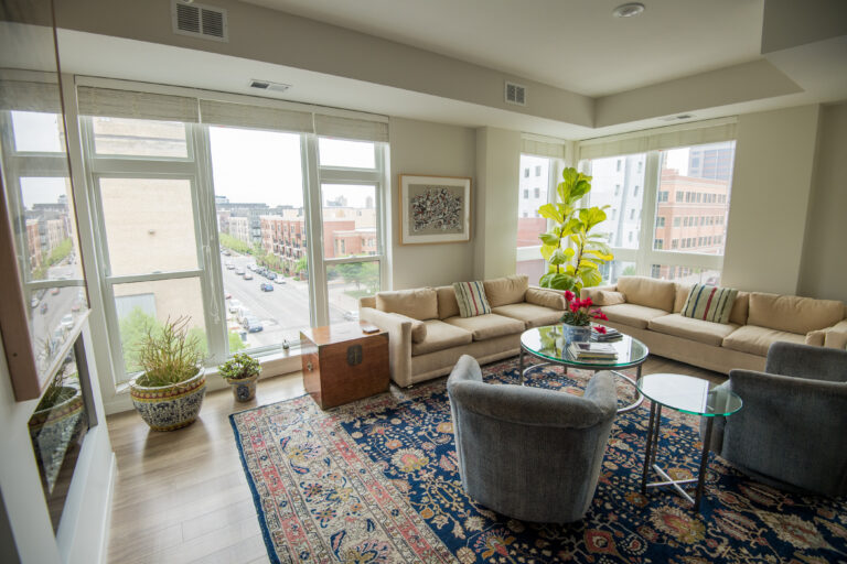 Modern living room with large windows, beige sofas, patterned rug, and city view outside.