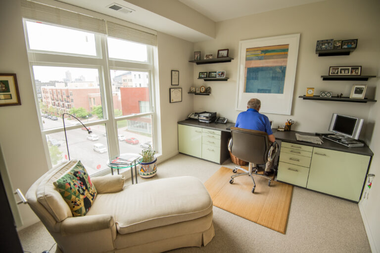 A man sits at a desk in a bright home office with large windows and modern decor.