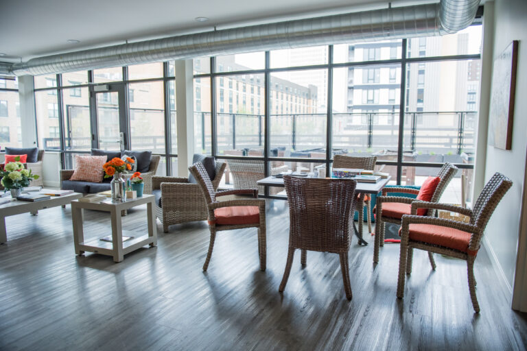 Bright, modern lounge with wicker chairs, sofas, flowers, and large windows overlooking city buildings.