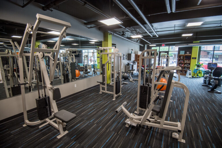 A modern gym with weight machines, cardio equipment, mirrors, and windows letting in natural light.