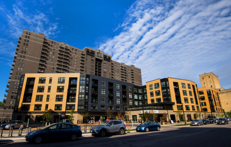 Modern apartment buildings with cars parked and driving on a street under a blue sky with scattered clouds.