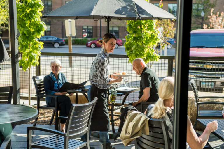 A smiling server brings food to outdoor diners at a sunny restaurant patio with green vines and black chairs.