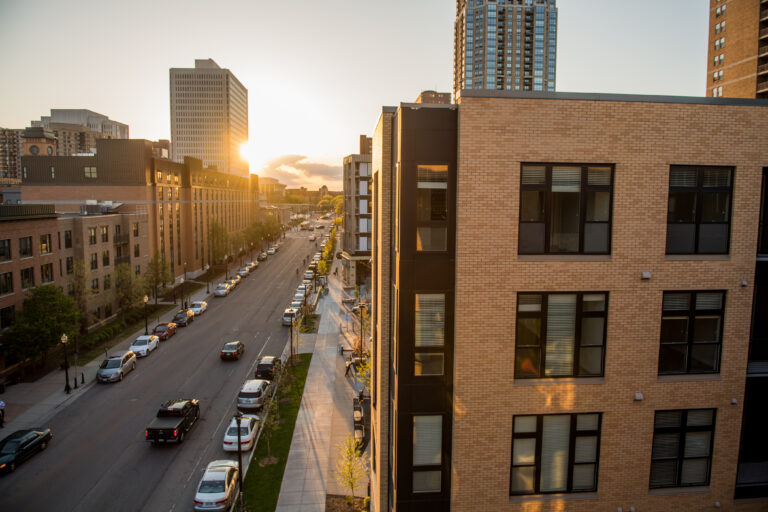 A city street at sunset with parked cars, modern buildings, and the sun setting behind tall skyscrapers.