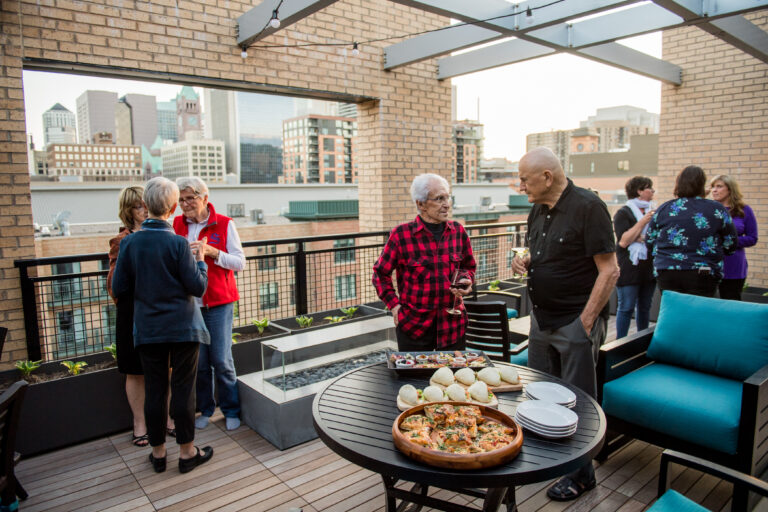 Older adults socializing on a rooftop patio with city views, food, and drinks on a table in the foreground.