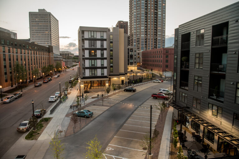 A modern city street with tall buildings, parked cars, and empty parking spaces at dusk.