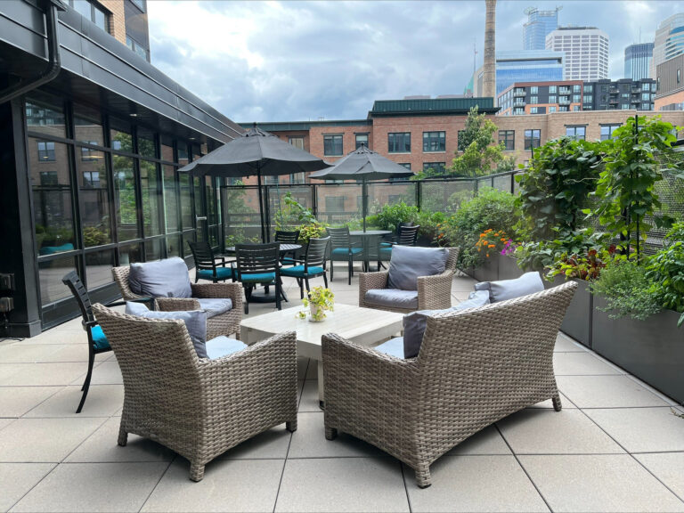 Modern rooftop patio with wicker chairs, tables, umbrellas, and potted plants, set against a city skyline.
