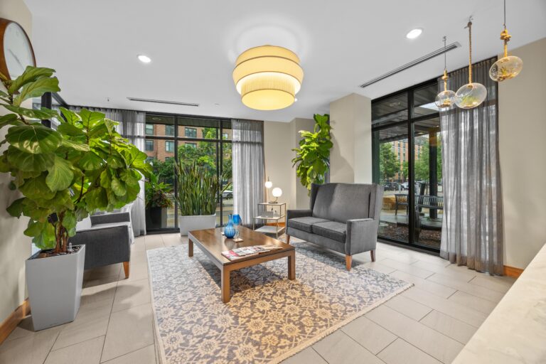 Modern lobby with gray sofas, potted plants, a coffee table, rug, and large windows letting in natural light.