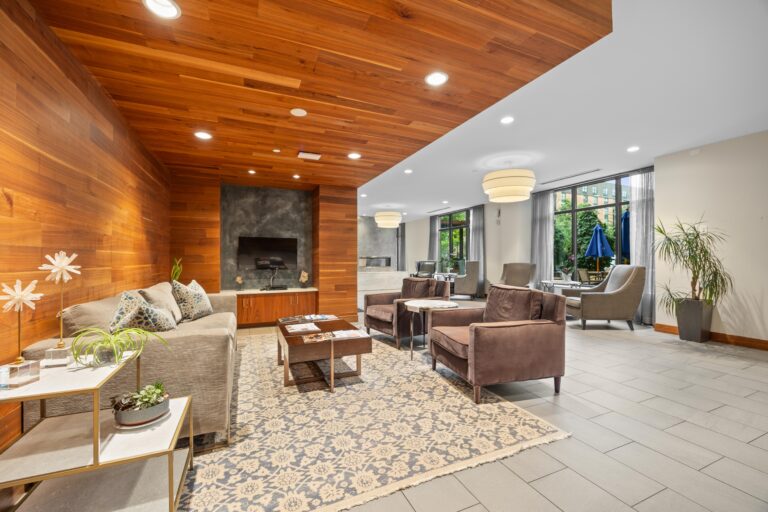 Modern lounge with wood-paneled ceiling, sofas, chairs, large rug, and windows overlooking greenery.