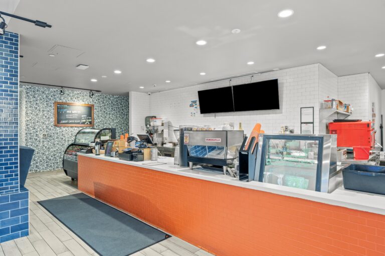 Modern coffee shop counter with coffee machines, display case, and blue and orange tile accents.