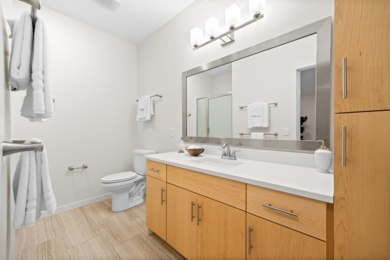 Modern bathroom with light wood cabinets, large mirror, white countertop, toilet, and towels on racks.