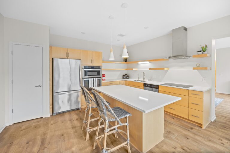 Modern kitchen with light wood cabinets, stainless steel appliances, and an island with three woven chairs.