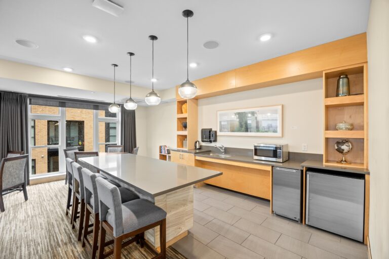 Modern kitchen with island seating, pendant lights, wood cabinets, appliances, and large windows.