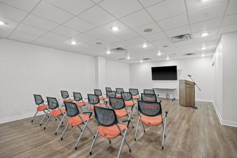 Small conference room with rows of orange chairs facing a podium, microphone, and large TV screen.