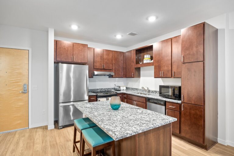 Modern kitchen with wood cabinets, granite island, stainless steel appliances, and two teal cushioned stools.