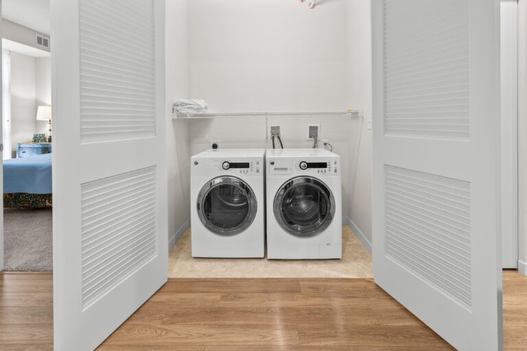Front-loading washer and dryer in a closet with open double doors and a shelf above them.