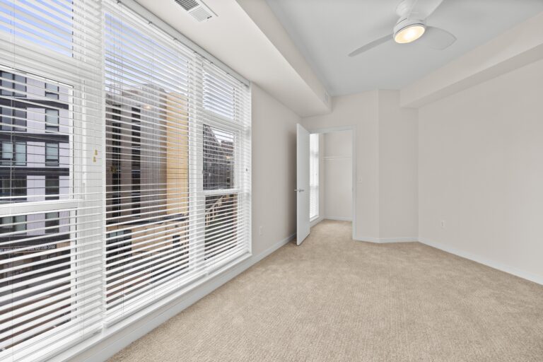 Bright, empty bedroom with beige carpet, large windows with blinds, ceiling fan, and open closet door.