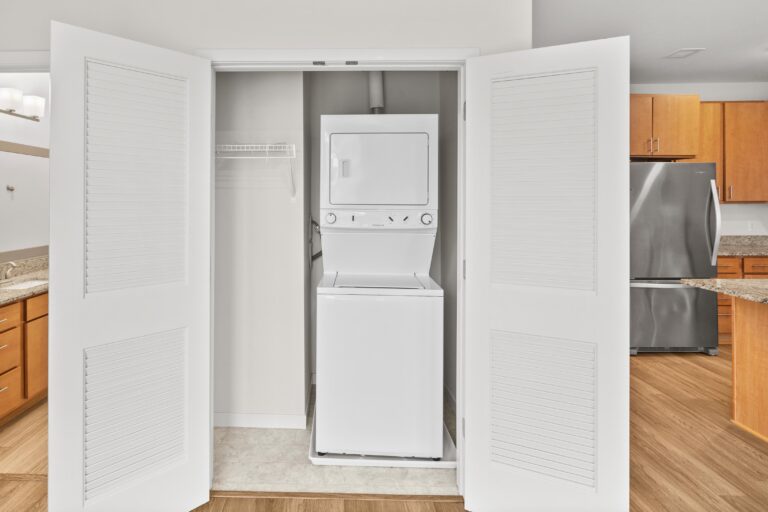 Stacked washer and dryer in a white closet next to a kitchen with wood cabinets and stainless steel appliances.