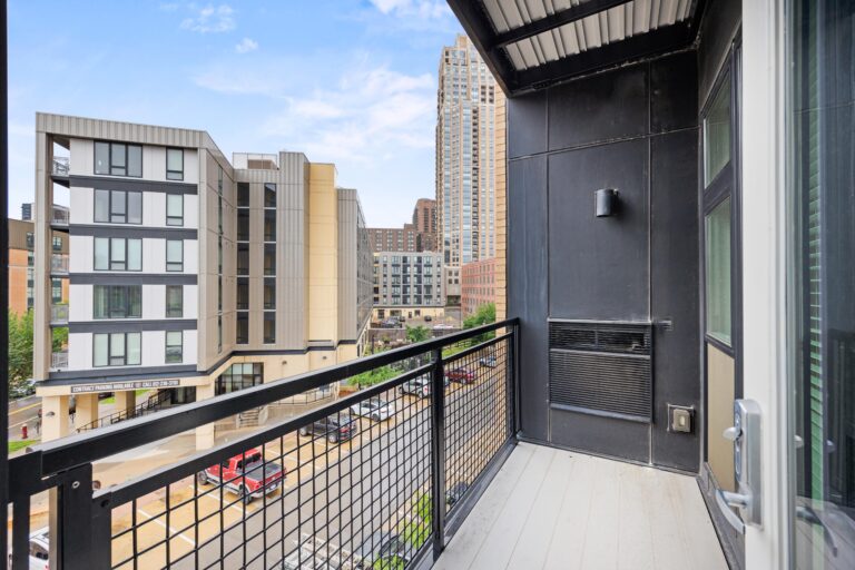 View from a modern balcony overlooking city buildings and a street with parked cars on a partly cloudy day.