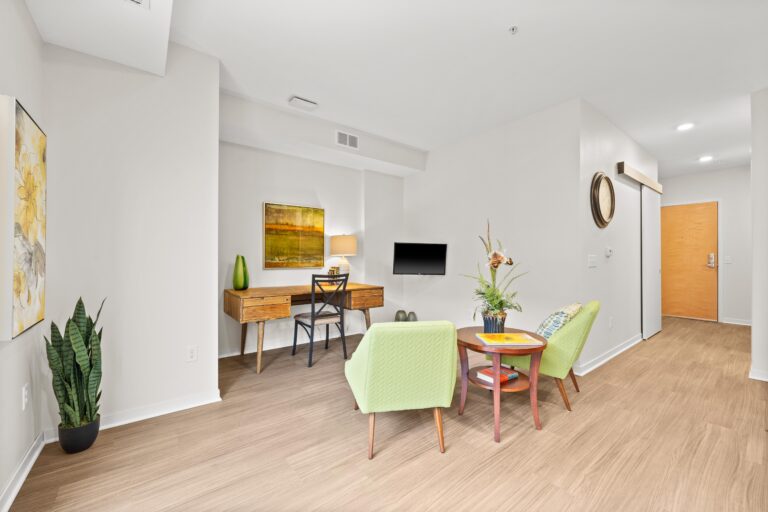 Modern living space with two green chairs, a small table, a desk, and a potted plant on light wood flooring.