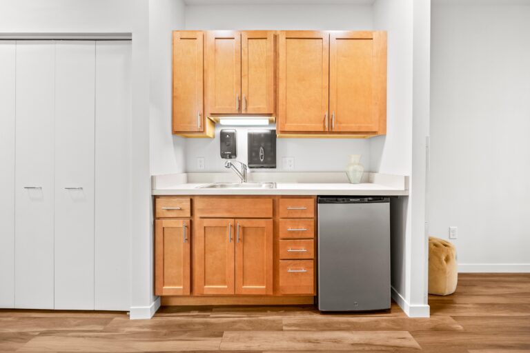 Compact kitchenette with wooden cabinets, a sink, dishwasher, and white countertop against a white wall.