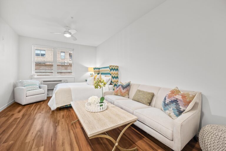 Bright studio room with a bed, sofa, armchair, coffee table, and large window; wood floors throughout.