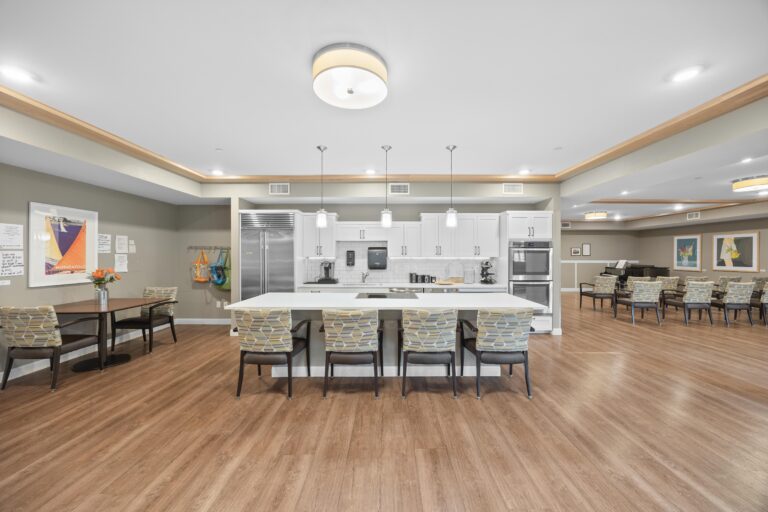 Modern communal kitchen with island seating, wooden floors, tables, and wall art in a spacious, bright room.