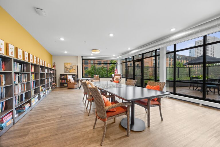 Modern library room with bookshelves, table and chairs, large windows, and cityscape view outside.