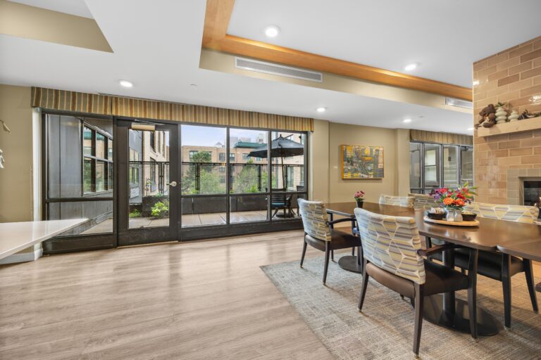 Modern dining room with large windows, outdoor patio view, and a table set for six with flowers.