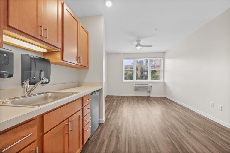 Modern kitchen with wood cabinets and open living area, featuring large window and wood-style flooring.