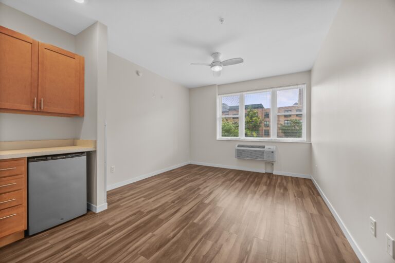 Bright, unfurnished room with wood flooring, large window, ceiling fan, and small kitchenette area.