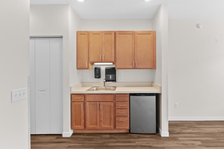 Small kitchenette with wooden cabinets, a sink, mini fridge, and light-colored countertops in a modern room.