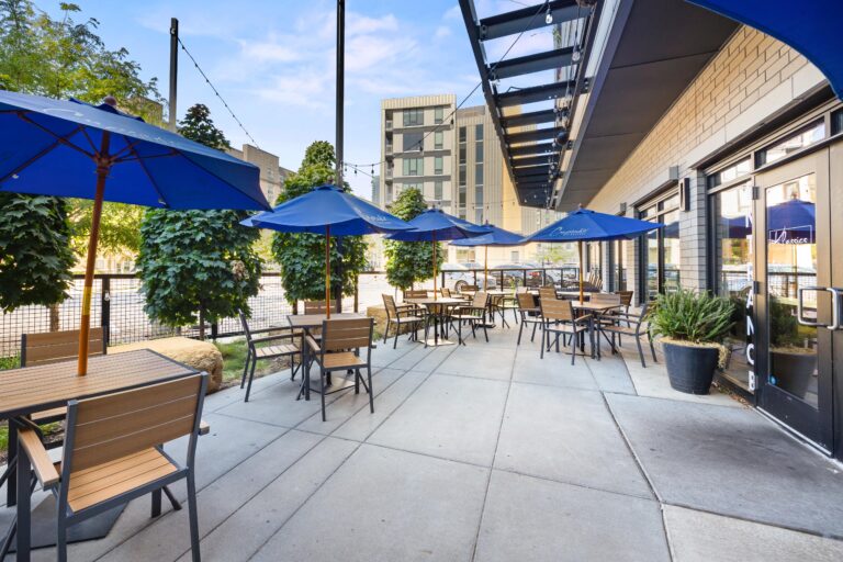 Outdoor café patio with wooden tables, chairs, and blue umbrellas on a sunny day in an urban setting.