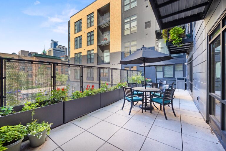 Modern apartment balcony with potted plants, outdoor dining set, and city buildings in the background.