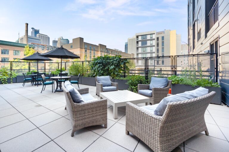 Outdoor patio with wicker lounge chairs, dining tables, umbrellas, and city buildings in the background.