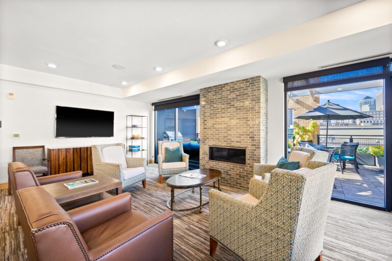 Modern lounge with armchairs, a TV, fireplace, and large windows opening to a sunny outdoor patio.