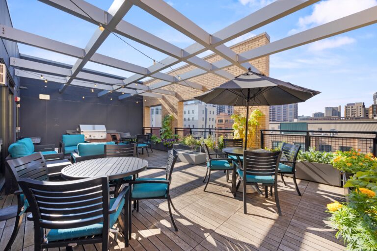 Rooftop patio with tables, chairs, a grill, umbrella, string lights, and city buildings in the background.