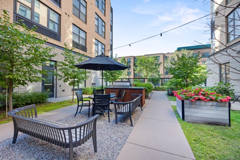 Outdoor patio with seating, table and umbrella, surrounded by greenery and apartment buildings.