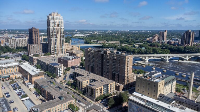 Aerial view of downtown Minneapolis with high-rise buildings, river, and a white bridge on a sunny day.