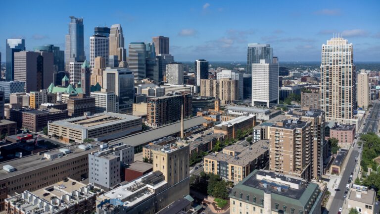 A city skyline with modern high-rise buildings under a bright blue sky with scattered clouds.