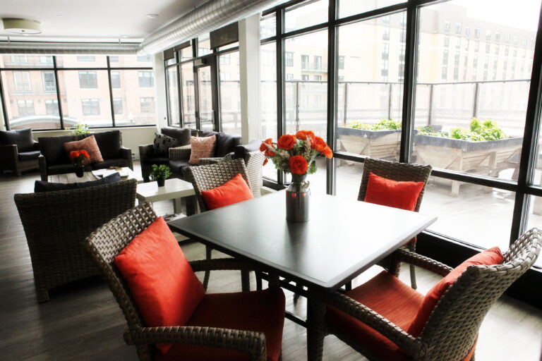 Modern lounge with wicker chairs, orange cushions, a table, and large windows overlooking a cityscape.