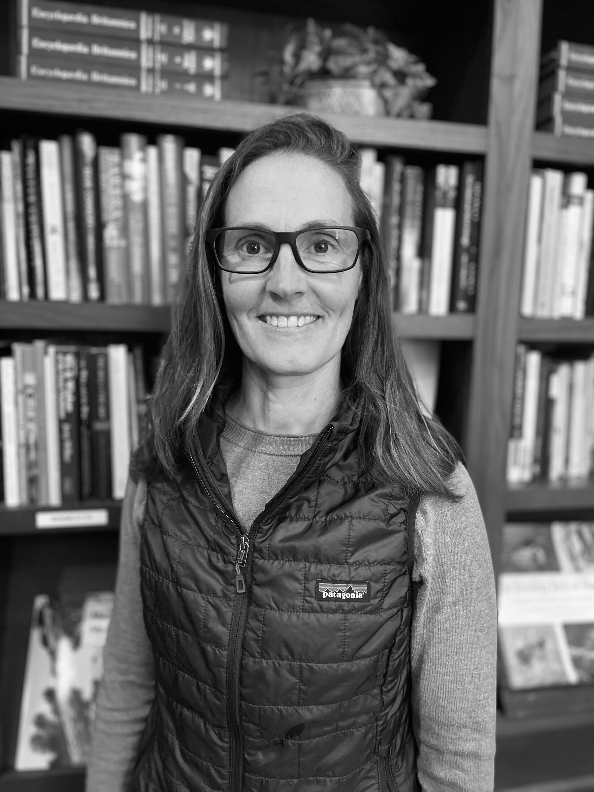 Portait of a woman with should-length dark hair and eye glasses, wearing a quilted outdoor vest and standing in front of a full book case. Black and white.