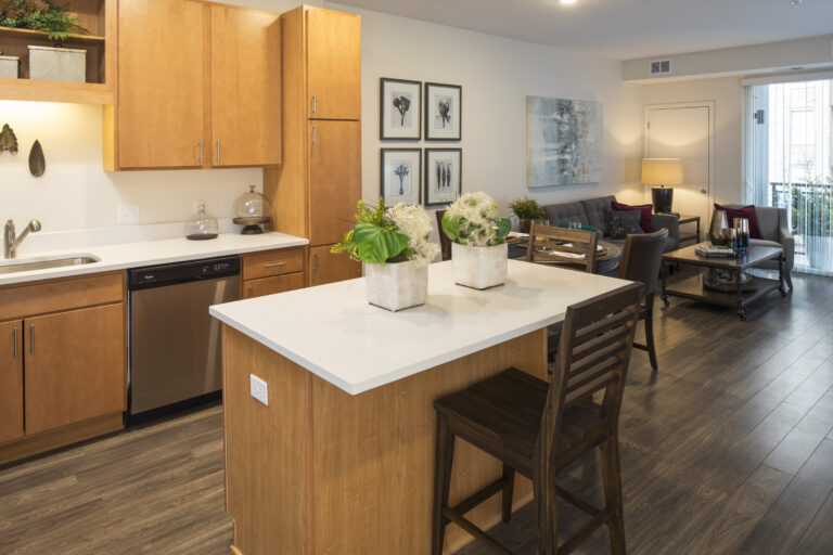 Modern kitchen with wooden cabinets, island seating, and a view of a cozy living room with decor.